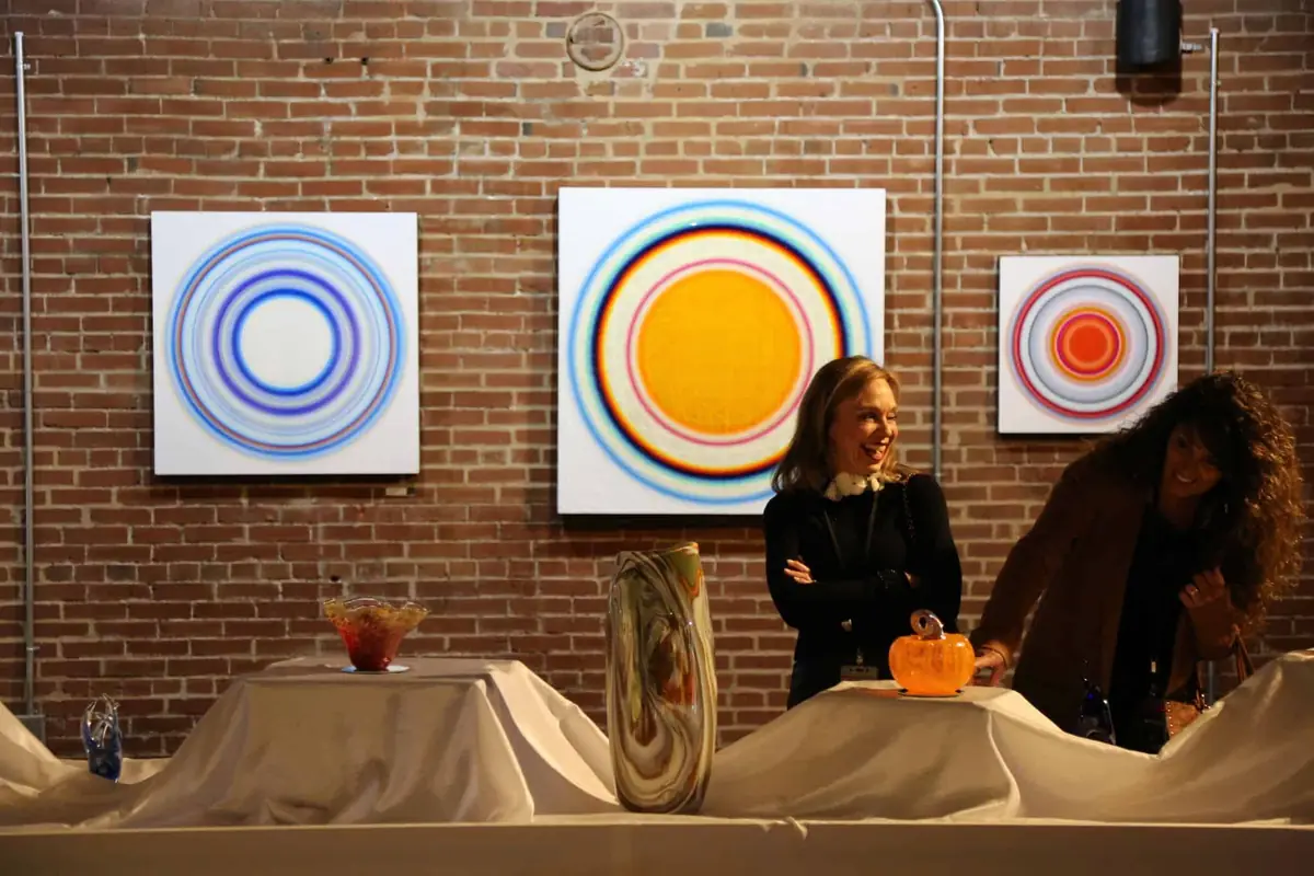 Guests talk beside hand-blown glass pieces on draped tables, with three circular paintings on a brick wall behind them.