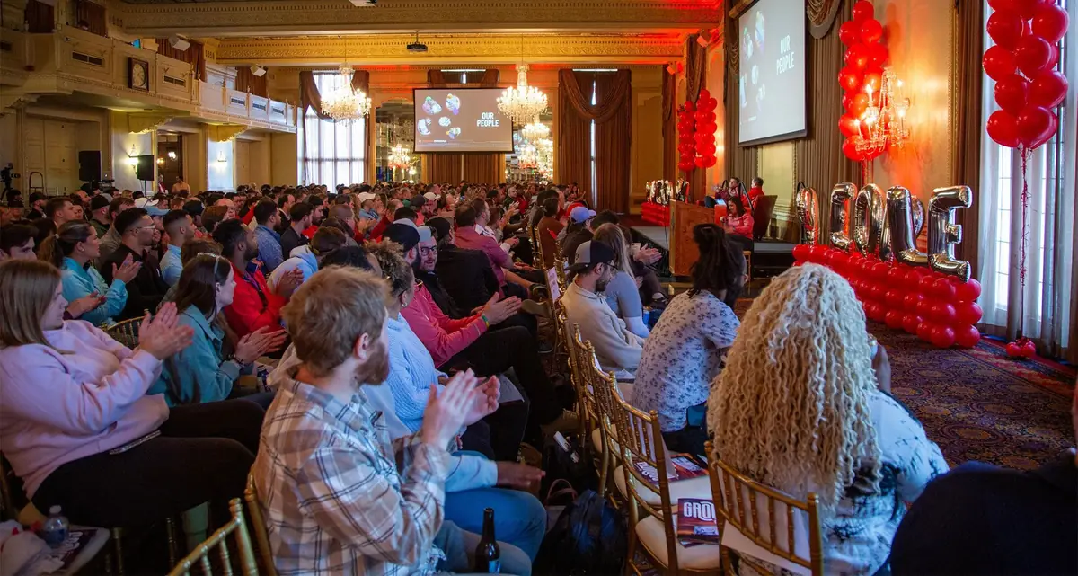 A ballroom full of Abstrakt team members applauding at a company meeting, with Our People on the screens